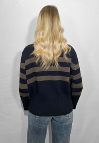 I capelli biondi e ondulati cadono su un maglione blu navy con strisce orizzontali marroni, abbinato a jeans blu sbiaditi, mentre si trova in piedi contro uno sfondo chiaro e semplice.