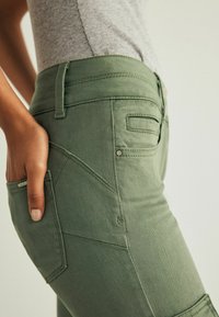 Kalhoty olivové barvy vyrobené z texturovaného denimu, se šikmými kapsami a decentním detailem loga. Design zahrnuje středně vysoký pasek.