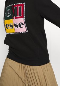 Černá mikina s kapucí s vícero barvami na přední straně, navržena ve čtvercích s vyšívanými detaily. Částečně viditelná plisovaná sukně v barvě pšenice.