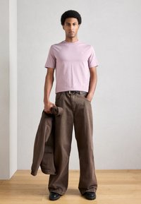 Νεαρός άνδρας που στέκεται σε εσωτερικό χώρο φορώντας ανοιχτό μωβ μπλουζάκι, καφέ παντελόνι φαρδιάς γραμμής, μαύρα παπούτσια, κρατώντας δίπλωτο καφέ σακάκι στο πλάι του.