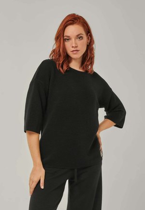 Junge Frau mit roten Haaren, die ein schwarzes, strukturiertes Halbarm-Oberteil und schwarze Hose trägt, posiert mit einer Hand auf der Hüfte und neutralem Gesichtsausdruck.