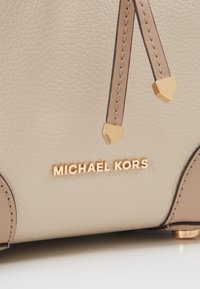 Gros plan d'un sac à main beige texturé avec un logo en métal doré et des sangles en cuir décoratives avec des embouts dorés.