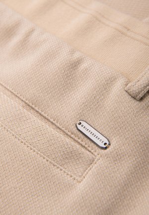 Béžová texturovaná látka s našitým detailem kapsy a malou kovovou štítkem označeným "QUIKTESSENCE" připevněným u okraje kapsy.