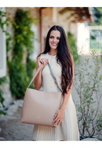 Beige Leder-Tote mit glatter Textur, ausgestattet mit einem verstellbaren grünen Trageriemen und dezentem Logo-Detail auf der Vorderseite.