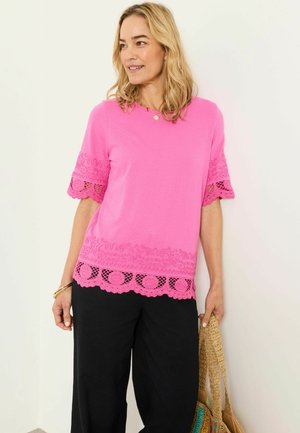 Vrouw draagt een felroze top met korte mouwen en kanten rand, zwarte broek, en houdt een gevlochten strozak vast tegen een witte achtergrond.