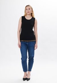 Top nero senza maniche con scollatura rotonda, caratterizzato da un accento circolare dorato sull'orlo, abbinato a jeans blu slim-fit e tacchi neri.