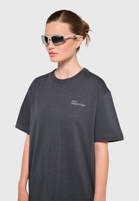 Okulary przeciwsłoneczne z ciemnymi szkłami i jasnoszarymi oprawkami, zestawione z luźną koszulką w kolorze grafitowym z białym napisem na piersi.