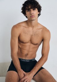 Schwarze Baumwollboxershorts mit einem weißen Calvin Klein-Logo am Bund, Modell sitzt vor einem hellen Hintergrund und zeigt eine durchtrainierte Figur.