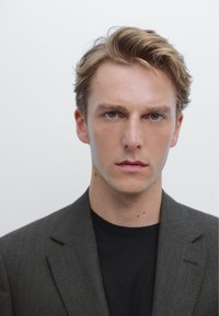 Ritratto di una persona con capelli castano chiaro, che indossa una giacca scura sopra una maglietta nera, con un'espressione neutra su uno sfondo bianco.