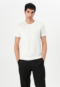 T-shirt de algodão branco com um decote redondo clássico e mangas curtas roladas, exibido em um modelo que veste calças pretas. Tecido liso e leve.