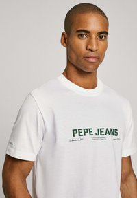 Balds kokvilnas t-krekls ar zaļu "PEPE JEANS" logo priekšpusē, apaļu kaklu un īsām piedurknēm, ar smalku gada norādi uz piedurknes.