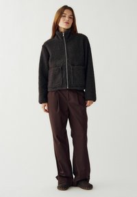 Donna che indossa una giacca in pile nera con cerniera e tasche frontali, pantaloni larghi marroni e scarpe marroni con borchie, in piedi contro uno sfondo neutro.