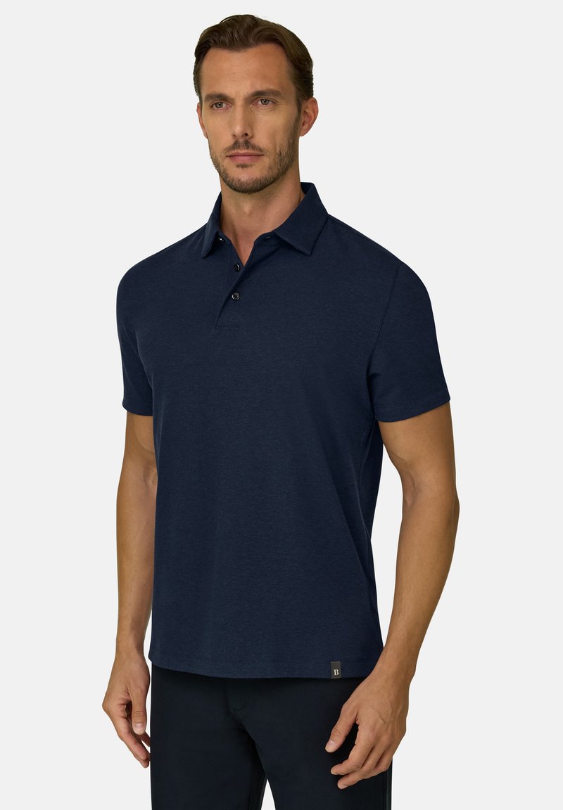 Homme portant un polo bleu foncé à manches courtes avec un col et trois boutons, debout devant un fond clair uni.