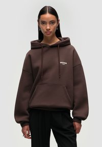 Brown oversized hoodie with a front pocket, drawstring hood, and small white logo on the chest. Soft, textured fabric with cuffed sleeves.