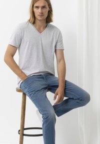T-shirt grigio a maniche corte con scollo a V abbinato a jeans in denim azzurro chiaro. L'uomo è seduto su uno sgabello di legno, con le mani nelle tasche.