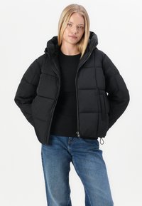 Chaqueta de plumas negra con cremallera frontal, capucha y diseño acolchado. Presenta bolsillos laterales y una cintura ajustada. Combinada con una camiseta negra y jeans azules.