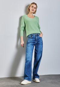 Maglione a maglia verde menta con scollo a V, abbinato a jeans blu a gamba larga e sneakers bianche, su uno sfondo grigio.
