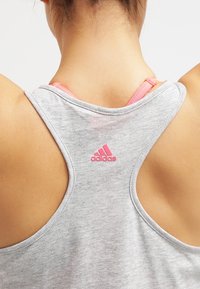 Šedý sportovní top s hlubokým výstřihem a designem racerback, se malým růžovým logem Adidas na zadní straně a viditelnými růžovými ramínky pod ním.