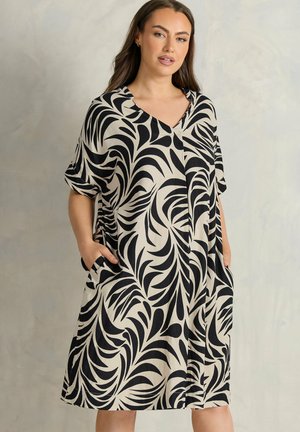 Frau trägt ein knielanges beiges Kleid mit schwarzem abstraktem Blattmuster, kurzen Ärmeln, V-Ausschnitt und Händen in den Seitentaschen.