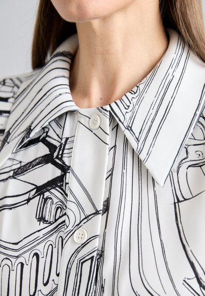 Blusa blanca con dibujos en línea negra, que presenta un cuello puntiagudo y un cierre de dos botones, textura suave y un diseño minimalista.