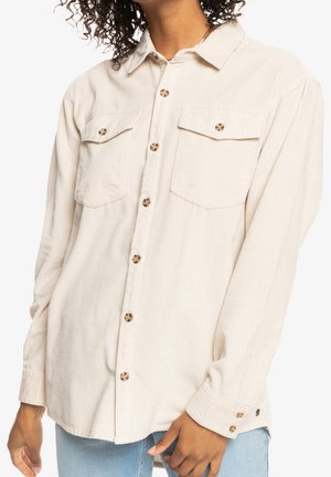Kvinde iført en cremefarvet kordfløjls-skjorte med brystlommer og knapper i skildpaddeskal, kombineret med lyseblå jeans.