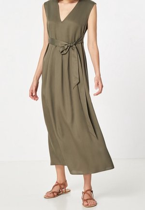 Olivgrünes, ärmelloses Maxikleid mit V-Ausschnitt, gebundenem Taillenband und fließendem Stoff. Kombiniert mit braunen flachen Sandalen.