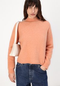 Pulover piersiciu, pufos, cu guler înalt și mâneci lungi, asortat cu blugi albaștri. O geantă mică, de culoare deschisă, atârnă de umăr.