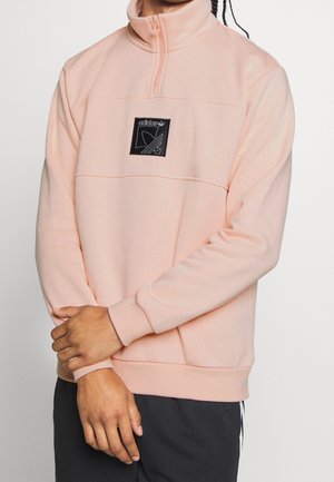 Personne portant un pull polaire Adidas rose clair avec une fermeture éclair au quart et un patch logo noir sur la poitrine, mains croisées devant.