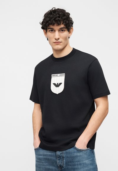 Νέος άνδρας με σγουρά μαλλιά που φοράει μαύρο μπλουζάκι Emporio Armani με λογότυπο ασπίδας στο μπροστινό μέρος και μπλε τζιν, με τα χέρια στις τσέπες.