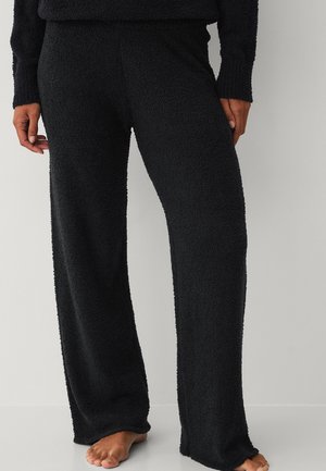 Persoon draagt zwarte, gestructureerde wijde broek met bijpassende top, staat op blote voeten op een lichte vloer tegen een effen muur.