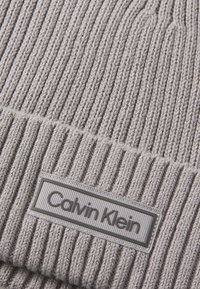 Gray ribbed knit beanie with a woven texture. It features a rectangular brand tag with "Calvin Klein" in embossed letters.