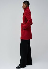 Crvena vuna kaput s visokim ovratnikom, bočnim džepovima, remenom u struku i dugim rukavima na dugmad, uparen s crnim širokim hlačama i cipelama s špicastim nožnim dijelom.