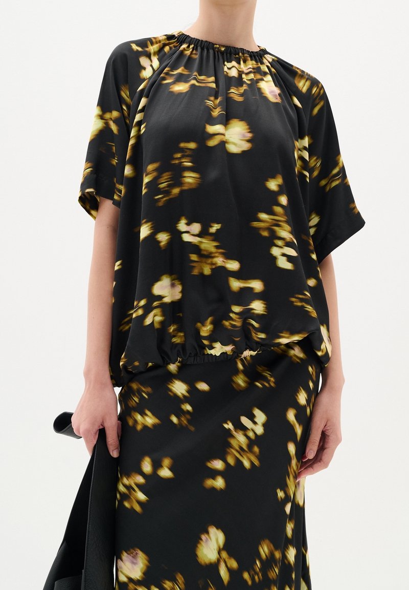 Blouse noire avec imprimé floral jaune, manches courtes, encolure froncée et ourlet élastique. Portée avec une jupe assortie, texture lisse.