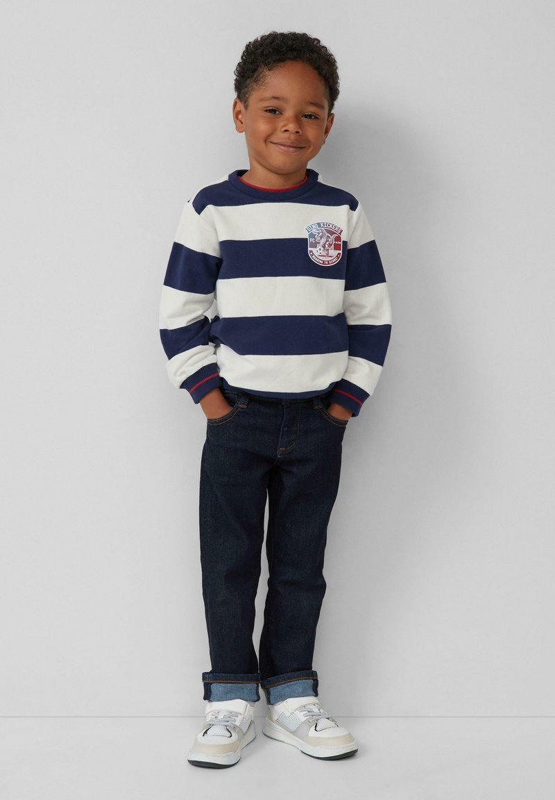 Maglietta a maniche lunghe a righe blu navy e bianche con un piccolo patch, abbinata a jeans in denim scuro e sneakers bianche. Design casual.