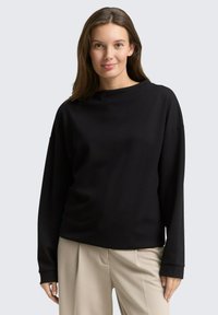 Haut noir à manches longues avec une coupe décontractée, un col rond et une texture côtelée. Associé à un pantalon large beige clair.