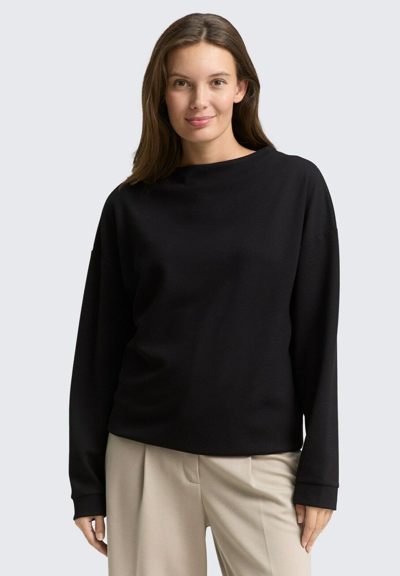 Haut noir à manches longues avec une coupe décontractée, un col rond et une texture côtelée. Associé à un pantalon large beige clair.