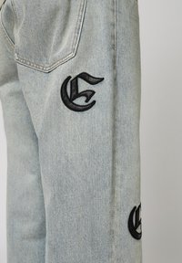 Vaqueros de mezclilla azul claro con logotipos bordados en negro en el lateral. Diseño clásico de cinco bolsillos con costuras visibles y una textura ligeramente desteñida.