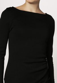 Černý top s dlouhým rukávem a lodním výstřihem, vyrobený z hladkého materiálu. Obsahuje nápadnou strukturu na jedné straně a minimalistické designové prvky.