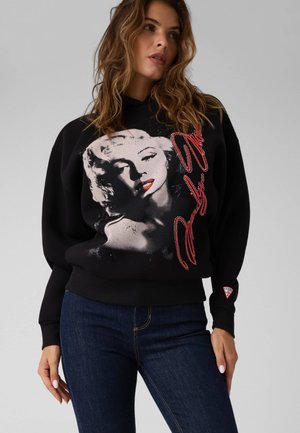 Kvinde iført sort sweatshirt med stort gråtonebillede af Marilyn Monroe og rødt kursiv tekst, kombineret med mørkeblå jeans.