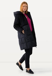 Lång svart pufferjacka med quiltad textur, överdimensionerad huva och dragkedjestängning. Parat med en rosa topp och slim fit svarta byxor. Glansiga skor.