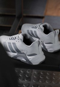 Paire de baskets blanches et grises Adidas placées sur un banc sombre avec un sol carrelé en arrière-plan.