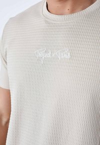 Camiseta texturizada de color beige claro con mangas cortas, que presenta un escote redondo y un logo bordado en blanco de "Project x Paris" en la parte frontal.