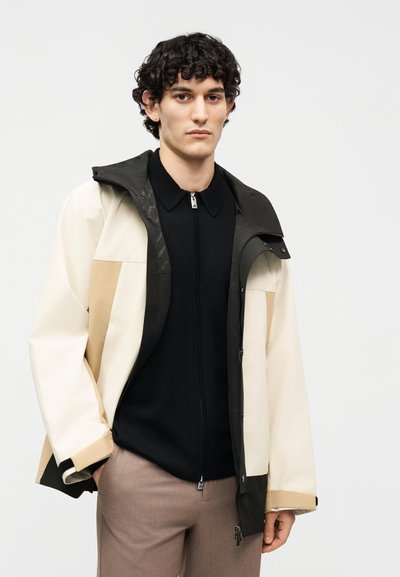 Νέος άνδρας με σγουρά μαλλιά φοράει ένα μπουφάν με χρωματικό μπλοκ σε μπεζ και μαύρο πάνω από μια μαύρη μπλούζα με φερμουάρ και καφέ παντελόνια, με φόντο λευκό.