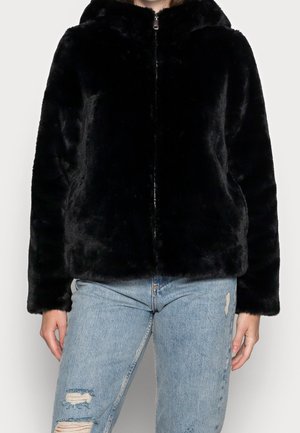 Persoon die een zwarte nepbonten jas met ritssluiting en capuchon draagt, samen met lichtblauwe geripte jeans, staand tegen een effen achtergrond.