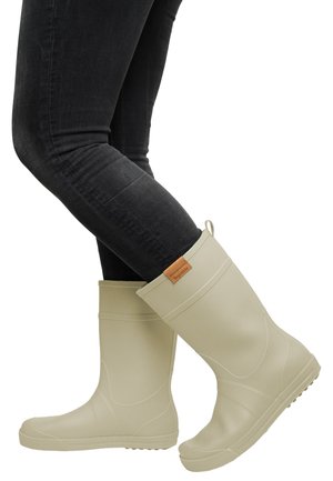 Person wearing beige rubber rain boots and black fitted jeans, standing with one foot slightly raised.