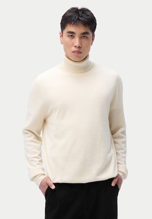 Junger Mann mit kurzen dunklen Haaren, trägt einen weißen Rollkragenpullover und schwarze Hosen, steht mit den Händen in den Taschen vor einem schlichten Hintergrund.
