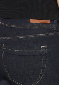 Jeans in denim blu scuro con dettagli di cucitura visibili, un'etichetta in pelle del marchio e tasche posteriori. Maglietta nera a coste parzialmente visibile.