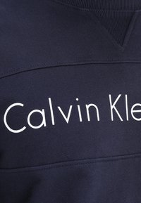 Felpa blu navy con una texture morbida che presenta il logo bianco "Calvin Klein". Il design include scollo a coste e dettagli cuciti.