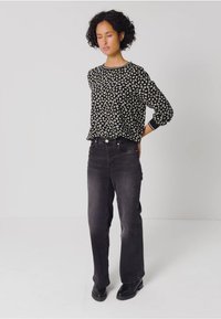 Schwarzes Langarmoberteil mit weißem abstraktem Muster, kombiniert mit dunklen hochgeschnittenen Jeans und schwarzen Stiefeletten, vor einem neutralen Hintergrund stehend.