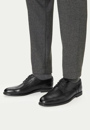 Schwarze Lederschuhe mit Schnürsenkeln, getragen mit grauen Socken und dunkelgrauen, maßgeschneiderten Hosen.